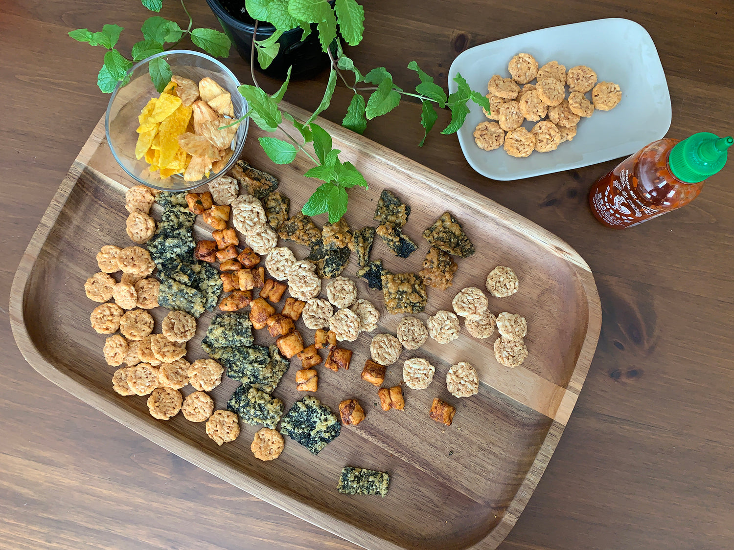 Top view of a snack board on a table by potted mint. Snackboard shows the many varieties of Woodridge Snacks and a bottle of Sriracha sauce.